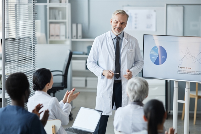 Man in lab coat next to a television with graphs on it. Presenting to a group of people clapping.